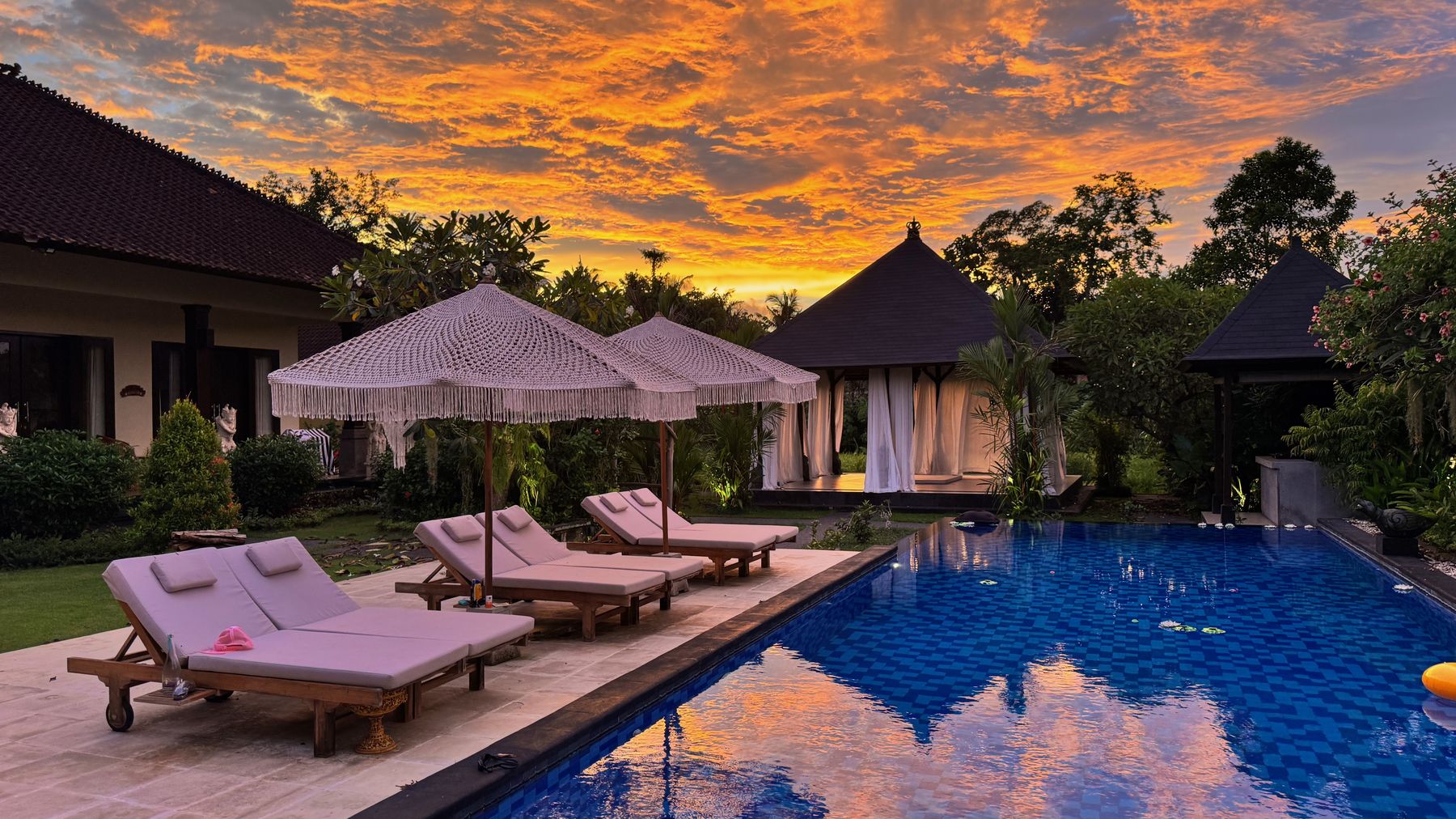 The pool at sunset, loungers along the edge and the sky on fire above the garden