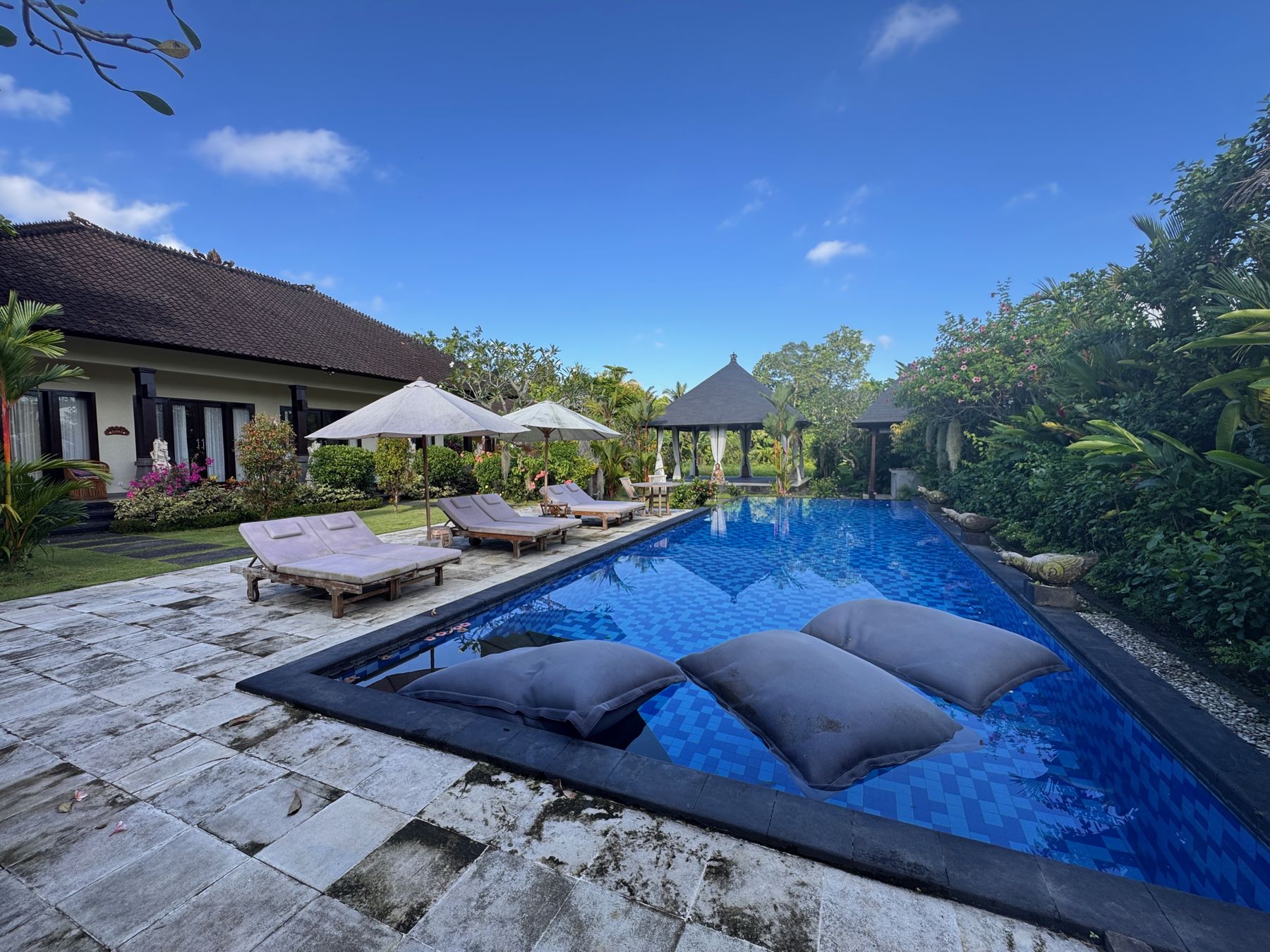 The pool on a clear day, loungers and umbrellas along the edge, cushions floating on the water