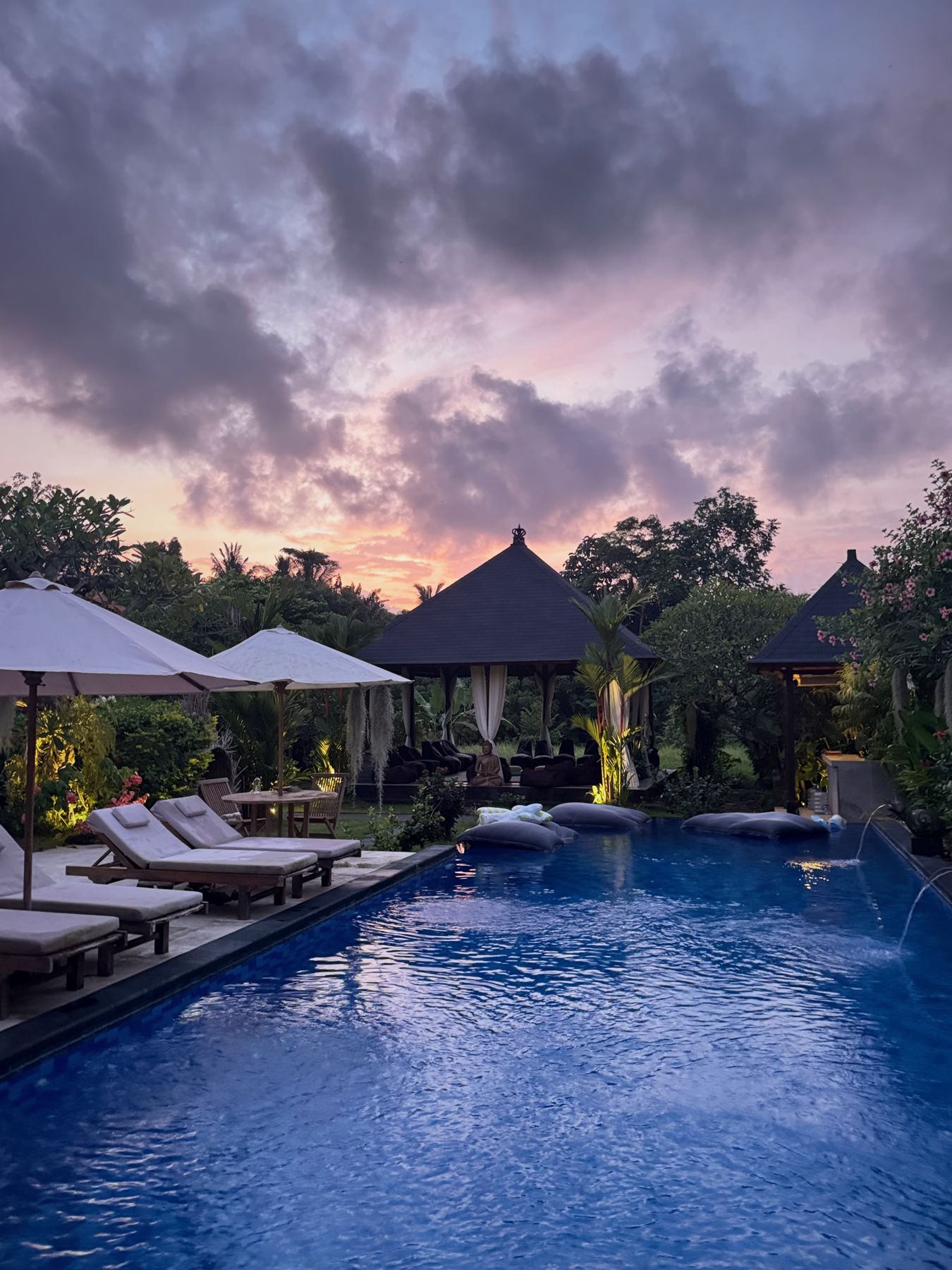 The pool at dusk under a violet sky, loungers along the edge and thatched roofs beyond